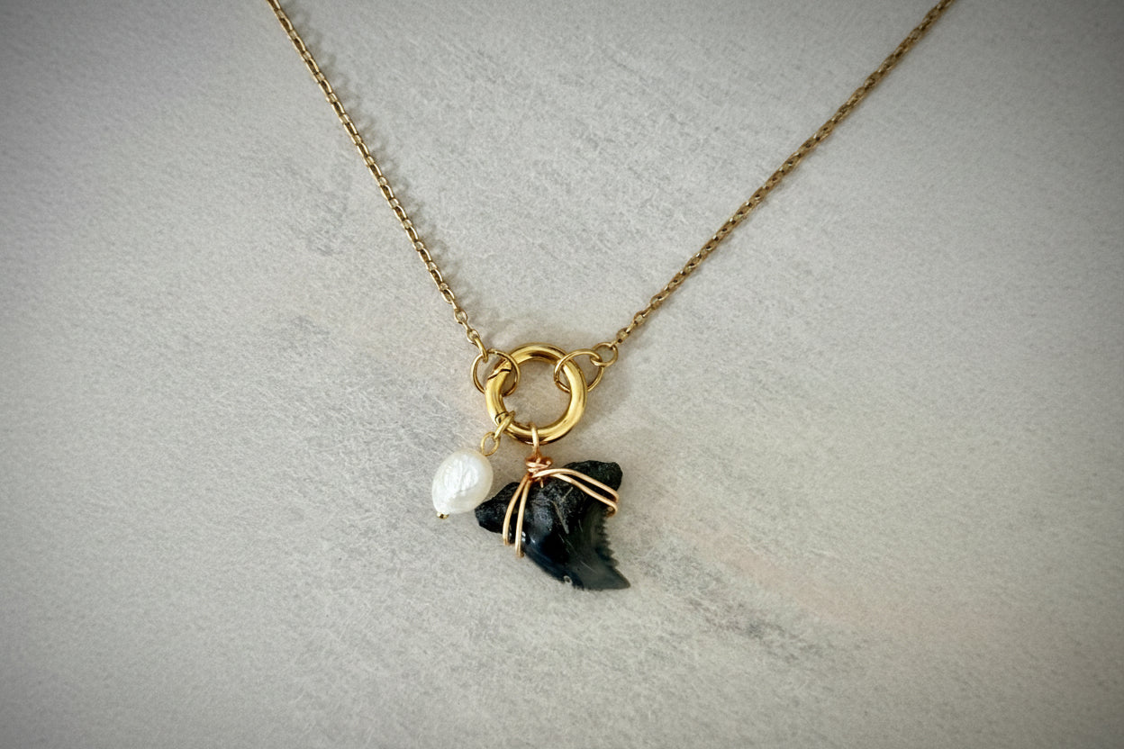 Necklace with gold ring, pearl, and black shark tooth pendant on a light gray background