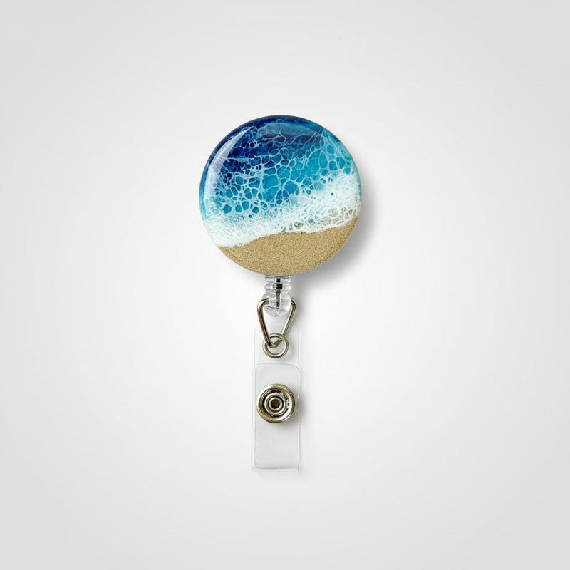 A round retractable badge reel with a beach-themed resin design showing blue ocean waves and white sand, attached to a clear plastic clip on a rustic wooden surface.