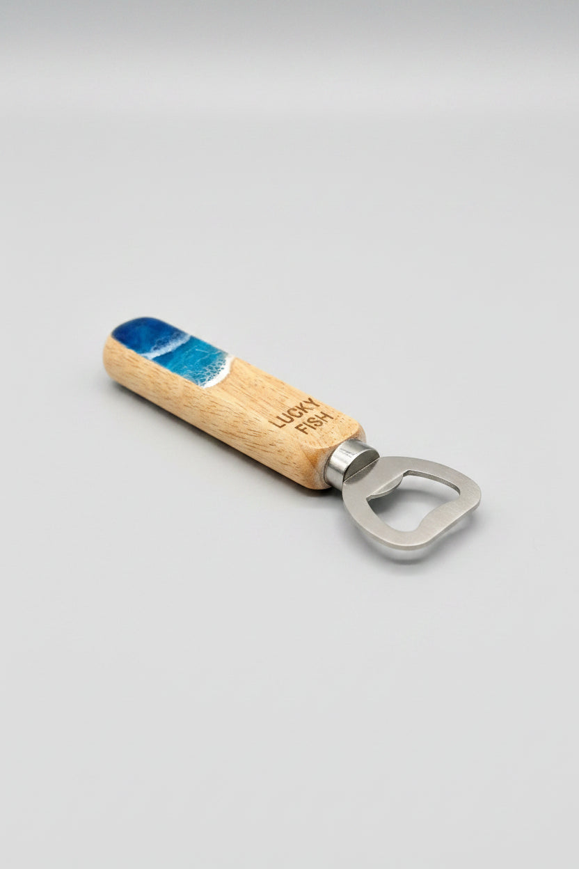 A wooden bottle opener with a blue and white resin wave-patterned handle and the engraved text 'LUCKY FISH', resting on a rustic wooden tabletop. Personalization available